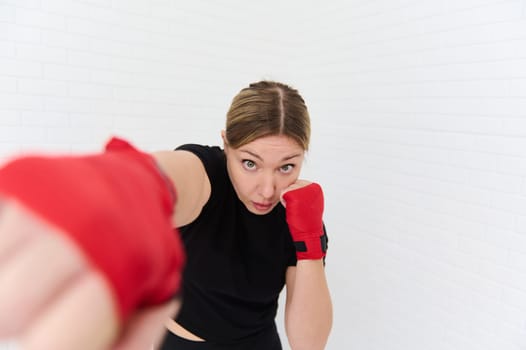 Caucasian strong athletic woman, boxer fighter with kickboxing red tapes on her hands, practicing boxing exercises, punching forward to camera. Martial art and combat. Wellness, activities and sports.