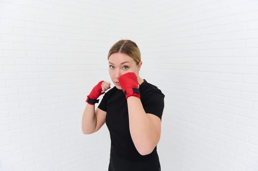 Authentic portrait of a young boxer woman, standing in self defense position, fighting during boxing training, dressed in black sportswear and wrapped wrists with red tapes, isolated white background