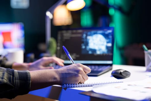 Male individual typing, and computing code for creating databases while writing on his notebook. Closeup of person working from home, monitoring software system and data center.