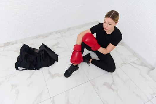 Overhead view of European young woman 30s, boxer fighter putting on red kickboxing leg pads guard. Sparring shin guards. Martial art, combat, challenge. Kickboxing concept. People, sport and fitness