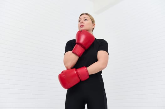 European confident woman boxer with red gloves and black sports wear, posing against white background. View from the bottom. Concept of female strength, combat training, and athletic discipline