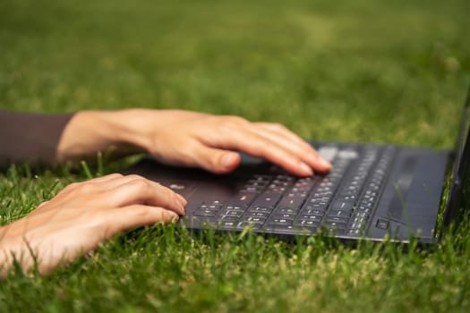 Female Hands are typing on the laptop keyboard, a young girl is engaged in freelancing, studies, resting on a meadow in the park, lying on the grass.