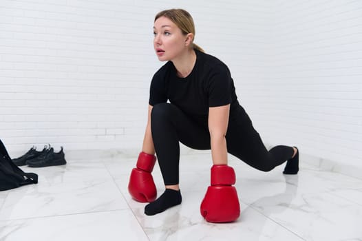 Woman boxer fighter stretching legs doing lunges, warming up her body before intensive boxing training, wearing red boxing gloves, exercising over white studio background. People and active lifestyle
