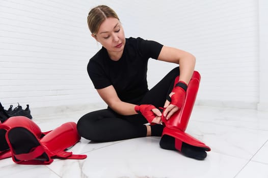Beautiful determines athletic woman boxer fighter putting on red kickboxing leg pads guard. Sparring shin guards, isolated over white background. Martial art, combat, challenge. Kickboxing concept.