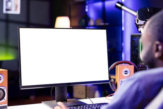 African american man in dimly lit apartment playing videogames on isolated screen gaming PC at computer desk, enjoying day off from work. BIPOC person enjoying game on mockup monitor