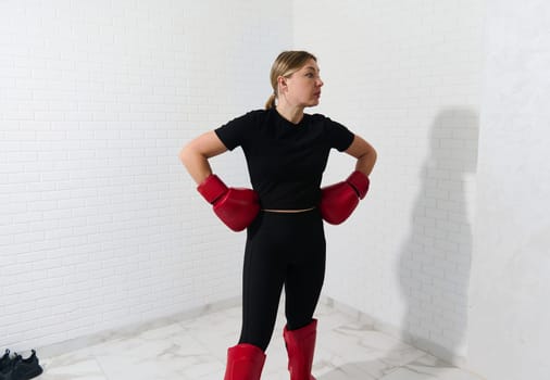 Confident woman boxer in boxing equipment, ready for kickboxing training, standing with arms on waist, isolated over white wall background. Martial art concept. Athletic discipline and Self defense
