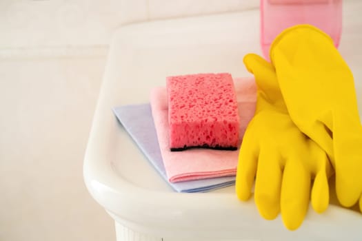 Household cleaning items, such as rubber yellow gloves, several types of rags and sponges, lie on a clean white washstand in the bathroom.