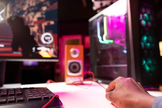 Gamer using keyboard and mouse to play FPS videogame on gaming PC tower with neon lit components. Man in apartment at night using high tech computer peripherals to defeat foes, close up