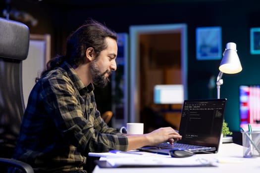 Male programmer works on his portable computer, managing the database and monitoring network for cyber security. Young man sitting at desk and using digital laptop for coding.