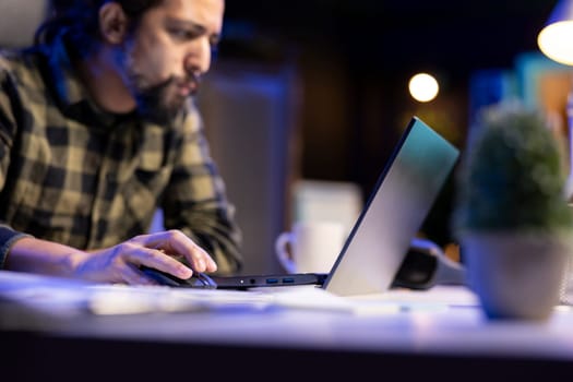 Closeup of man typing on laptop sitting in living room late at night and working. Serious male blogger using modern technology network wireless doing overtime for writing researching.