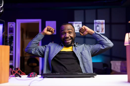 Portrait of delighted BIPOC man on desk chair celebrating winning videogame level in apartment. Happy african american player celebrating after achieving victory in game on gaming PC