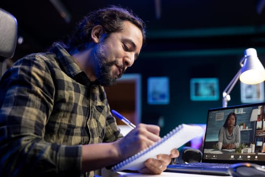 Closeup of bearded man with a pen and notebook, writing meeting notes while on video call on the laptop. Male individual positively working from home, using his notepad and portable computer.