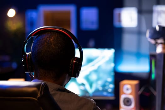 Focus on man playing videogames on computer monitor in blurry background, talking into headset. African american gamer using headphones to chat with his Esports team, close up
