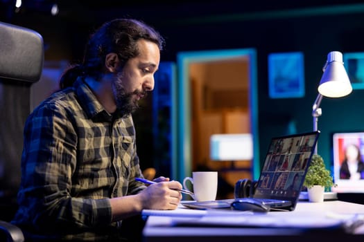 Working from home, a young man reviews his notes while speaking with a variety of people via video call. Serious male individual checks his notebook while attending a virtual meeting on his laptop.