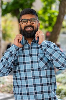 Portrait of happy smiling Indian bearded man listening music taking off headphones looking at camera, relaxation feel satisfied good news outdoors. Hispanic young guy on urban city street. Vertical
