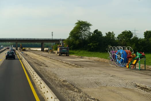 Weimar, Germany - May 7, 2023: Highway construction workers are seen using machinery while vehicles pass by. The focus is on road construction work near a scenic green area with an overpass in the background.