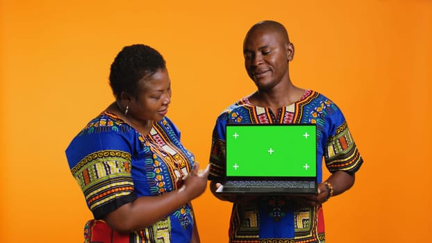 African american partners holding laptop with greenscreen on camera, presenting isolated display with chromakey and standing over orange background. Man and woman showing blank mockup.
