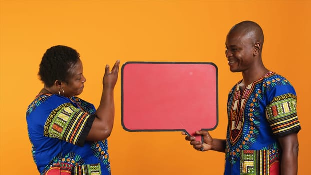 Married ethnic people pointing at red speech bubble icon made from cardboard for web promotions and advertisements. Young man and woman promoting something with isolated billboard sign.