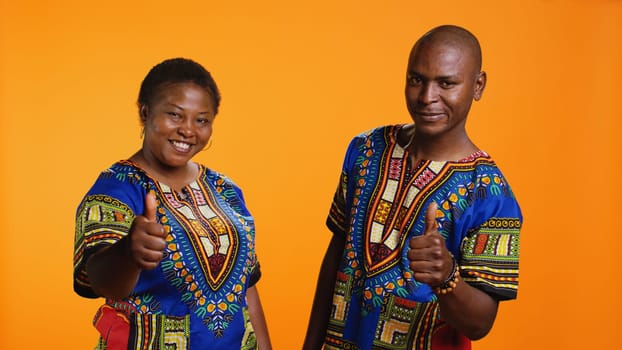 African american partners giving thumbs up gesture in studio, feeling cheerful and presenting their positive feedback. Married couple showing approval and doing like symbol on camera.