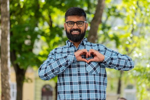 I love you. Indian young man makes symbol of love, showing heart sign to camera express romantic feelings, express sincere positive feelings outdoors. Charity, gratitude, donation. on city street