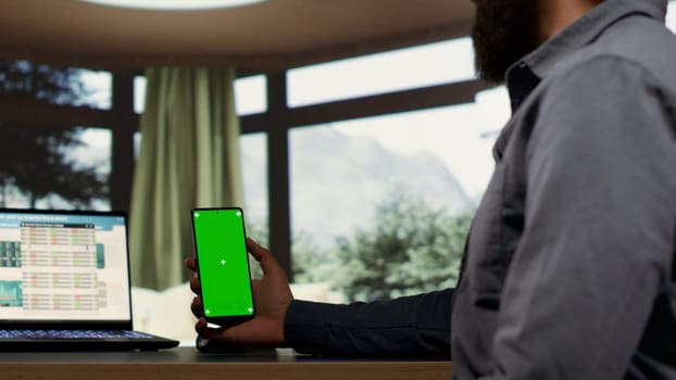 Entrepreneur holding smartphone with greenscreen template while he is examining global revenue trends on laptop. Company CEO connecting on device to help employees, using isolated copyspace.