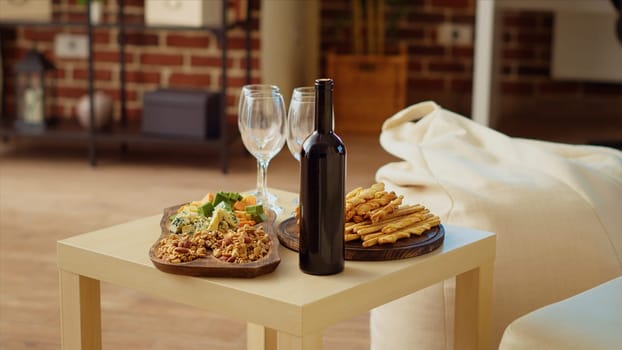 Panning shot of appetizers board and gourmet cheese platter set up by hosts to properly welcome guests arriving soon at social gathering. Wine bottle and glasses next to food in cozy home, close up