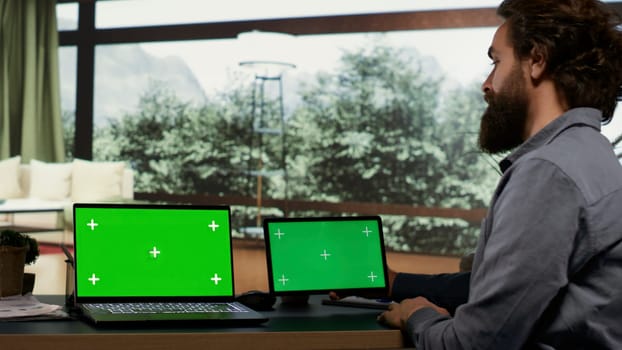 Wealthy person using greenscreen on two devices, sitting at his workstation in his modern elegant vacation home. Rich business owner and investor looking at screens running blank chromakey.