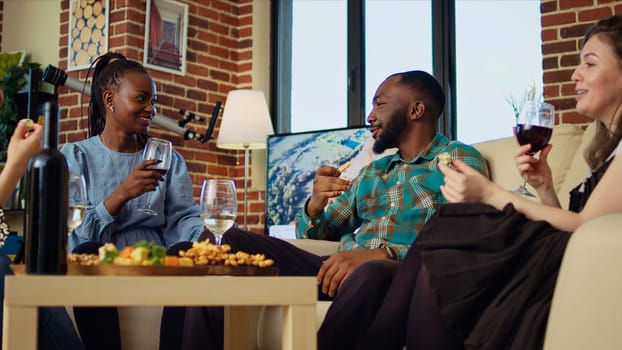 Interracial couple at social gathering talking on couch while enjoying glass of wine. Husband and wife guests at apartment party having pleasant conversation in brick wall home, eating snacks