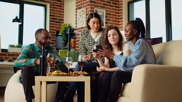 Friends at house party using smartphone to laugh at social media posts made by acquaintance while eating snacks and drinking wine. Guests using phone to look at images online, enjoying appetizers