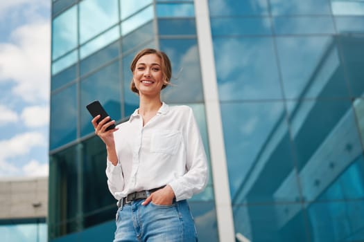 From below of smiling young businesswoman in smart casual holding smartphone while standing against modern office building. Confident entrepreneur texting via cellphone in city.