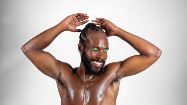 Portrait of confident shirtless, muscular African American gay man with green eyeshadow. Bearded man smiling with hands raised on white background. Happy gay individual with vibrant makeup.