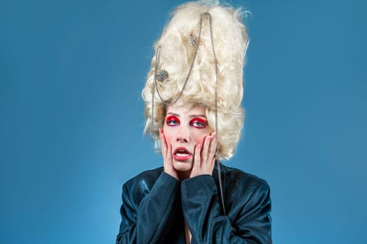 Portrait of silly princess wearing old-fashioned wig and red makeup, making funny faces. Funny woman in stylish jacket against blue background, like a royal party in blue studio.