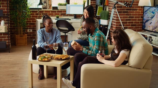 Multiethnic mates invited to birthday party offering presents to cheerful African american man impressed by nice gesture. Guests surprising their friend with thoughtful present and enjoying appetizers
