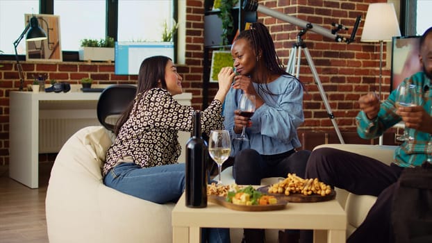 Happy women chatting on cozy couch at apartment party, drinking wine and enjoying conversation. Cheerful friends catching up with each other at home in stylish living room, talking