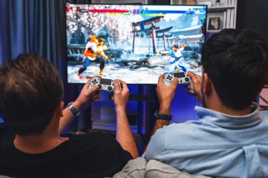 Friend gamers playing video game of battle martial arts fighter on TV using joysticks position of backside background. Comfy living neon light at home place with cheerful fighting winner. Sellable.