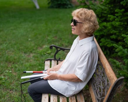 An elderly blind woman sits on a bench in the park with a folded tactile cane in her hands