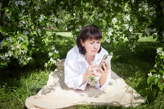 Brunette girl Using Smartphone in Blossoming Orchard in Springtime. Middle aged Woman enjoying phone among spring blossoms of apple or sakura trees