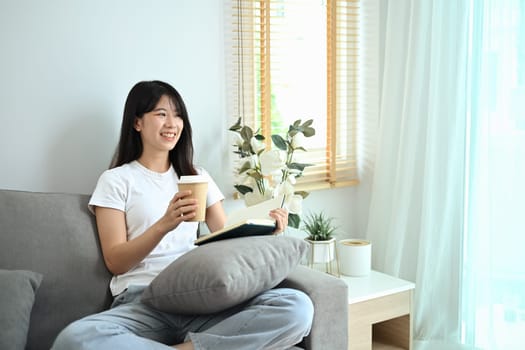 Charming young asian woman drinking coffee reading book on couch at home.