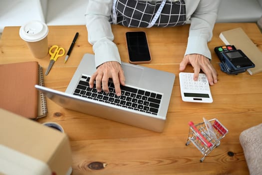 Above view of small business entrepreneur using laptop and calculating expenses at desk.