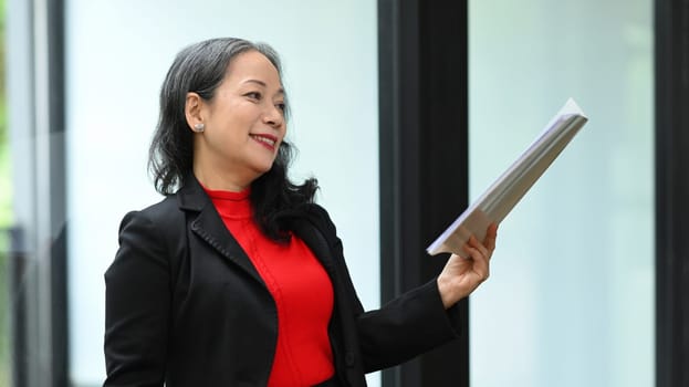 Confident mature businesswoman with financial document standing at workplace.