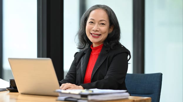 Smiling professional senior businesswoman sitting at the desk in front of laptop, looking at camera.