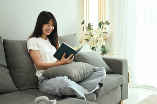 Smiling young woman sitting on cozy sofa and reading book. People, leisure and lifestyle concept.