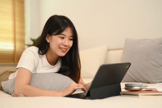 An attractive young Asian woman using digital tablet while lying on her bed.