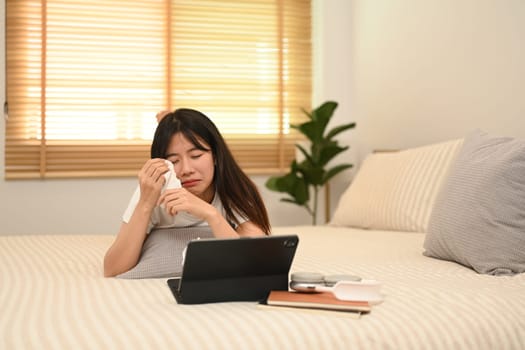 Sentimental young woman crying and wiping tears while watching dramatic movie in bed.