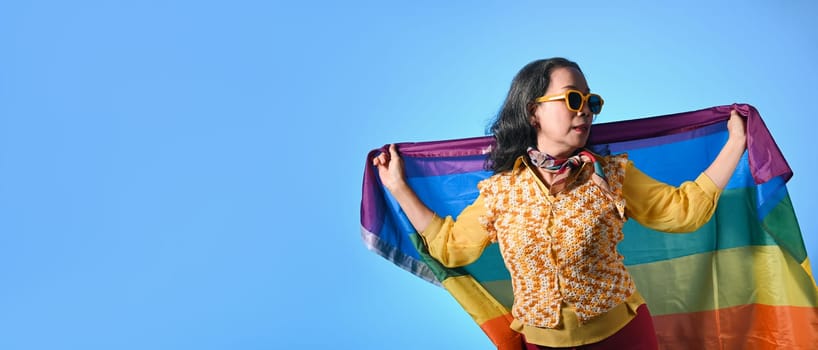 Beautiful mature woman waving rainbow flag on blue background. LGBT, human rights and equality concept.