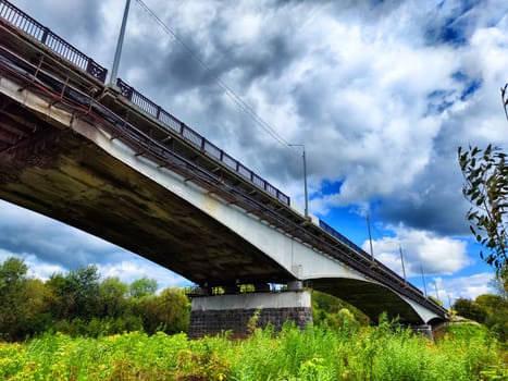 A large bridge extends over a calm river, with an overcast sky and urban buildings visible in the background