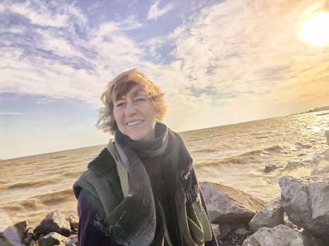 Happy cheerful middle aged woman in scarf taking selfie on nature outdoors and sun with water and waves of sea on the background