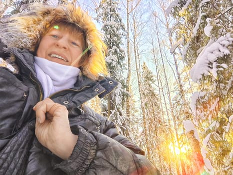 A cheerful middle aged woman in a winter coat with fur, scarf taking selfie outdoors on nature in a forest or park in beautiful sunny day