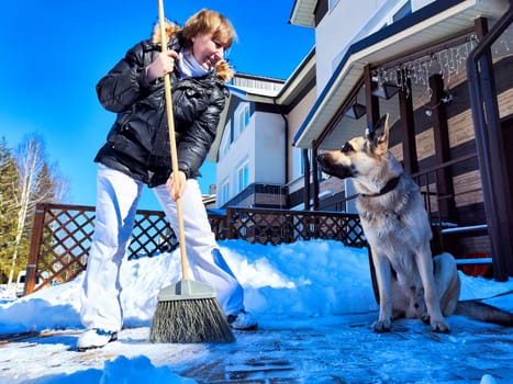 A girl or a woman with a snow broom at home and a large German Shepherd dog. Woman Clearing Snow at Home With Her German Shepherd