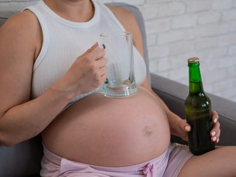 Faceless pregnant woman sitting on the sofa and holding a bottle of beer and a glass